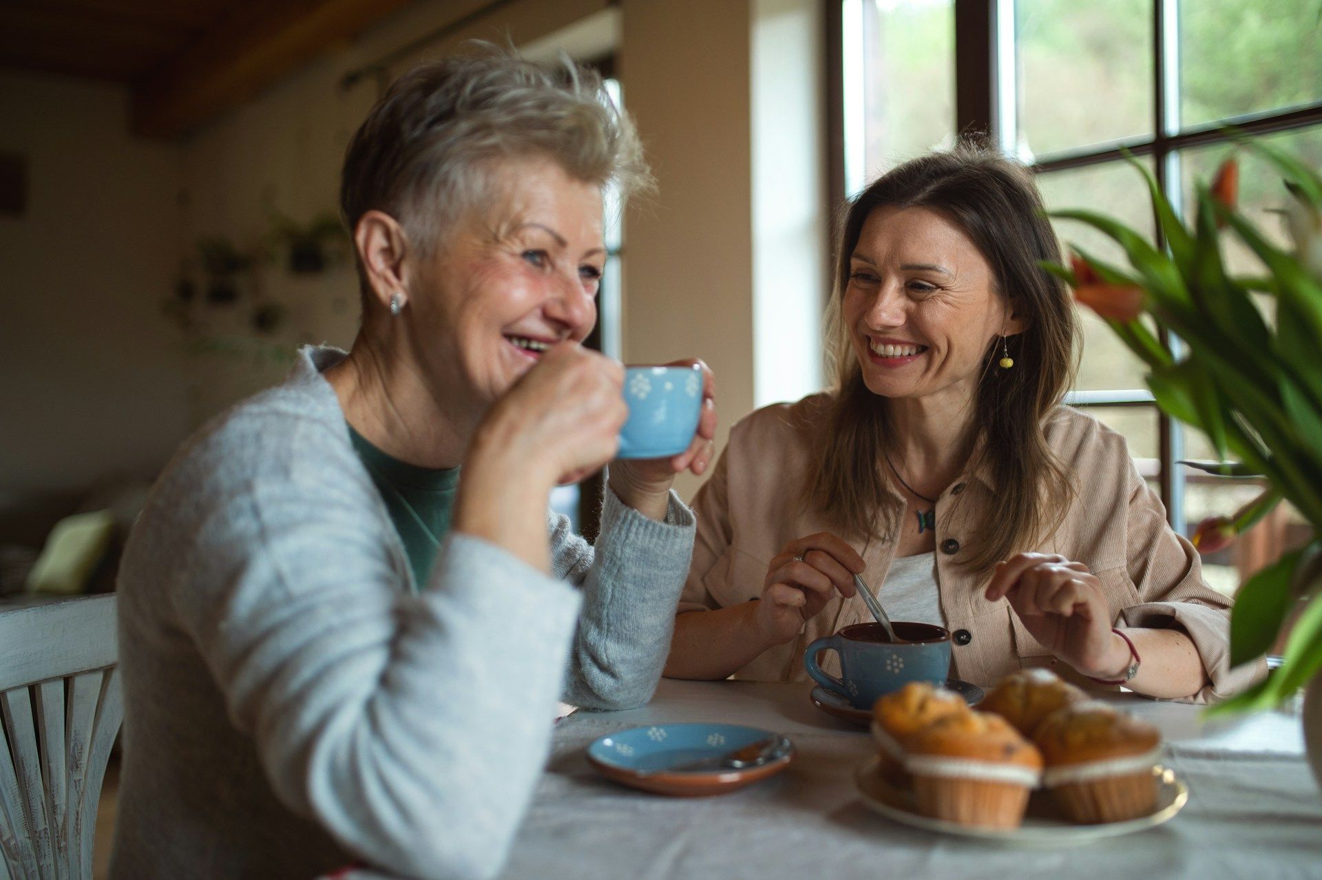 Two women smiling, drinking tea, and eating muffins at a table indoors.