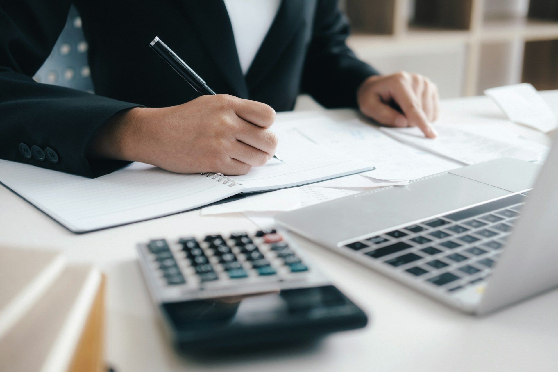 Person in a suit writing on paperwork, calculator, laptop, and books on a white desk.