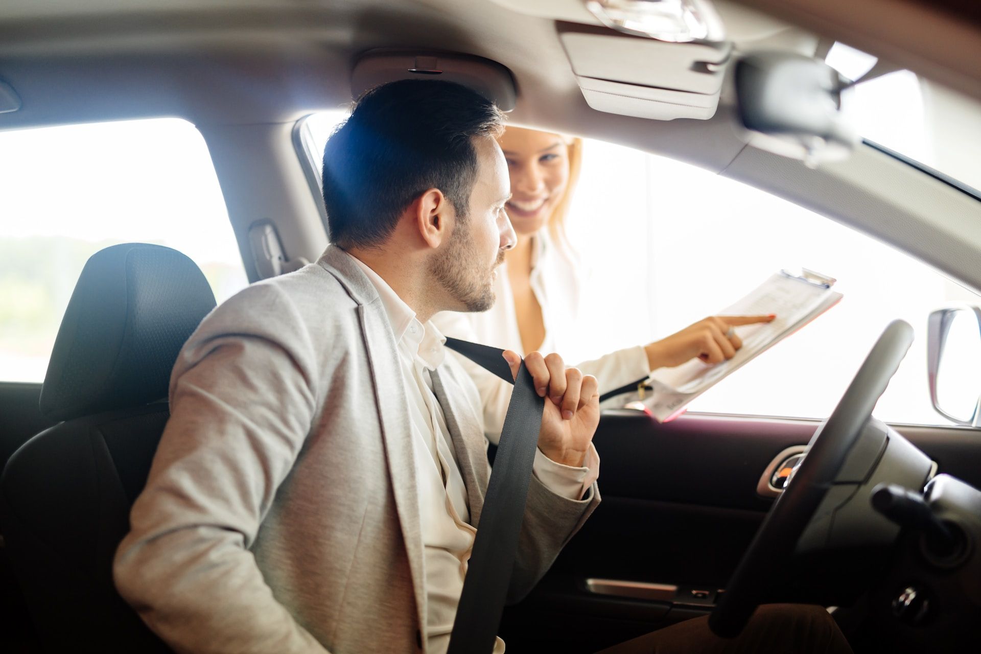 Man in car, woman pointing at paperwork. Car dealership setting.