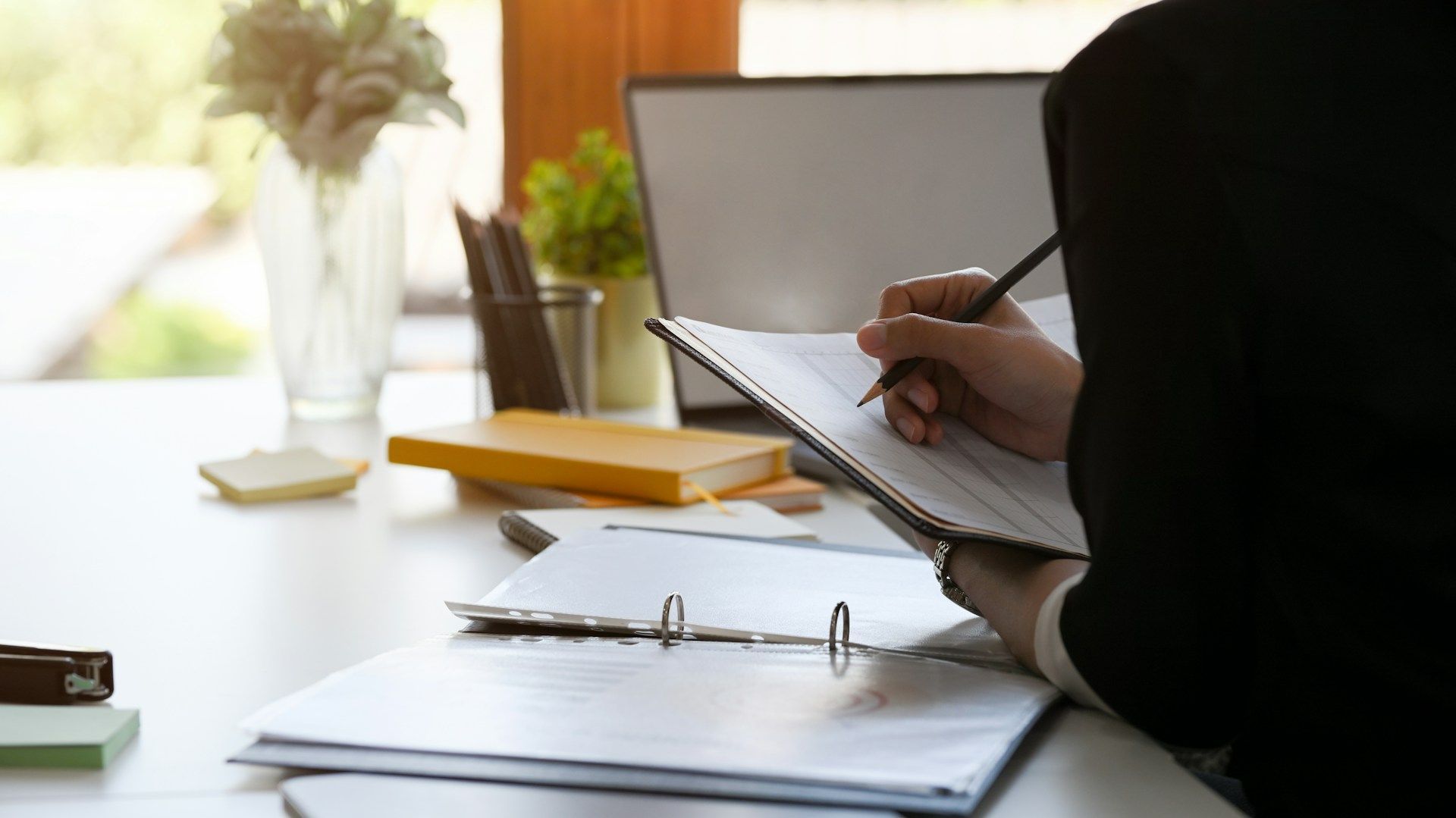 Person in a suit writing on a clipboard at a desk with papers, a laptop, and plants.