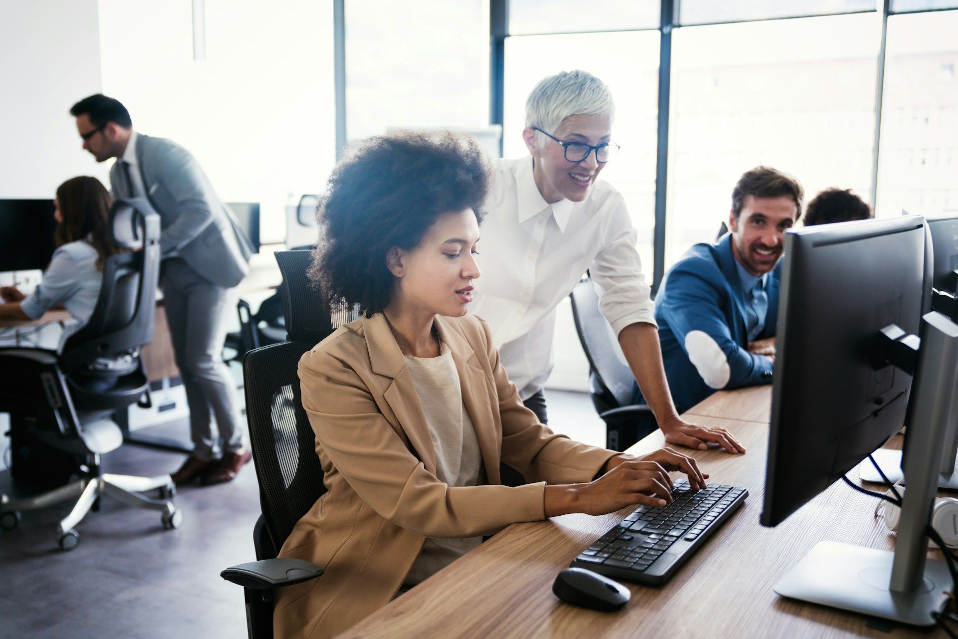 Office scene; woman working at a computer with a supervisor looking on; others work at desks.
