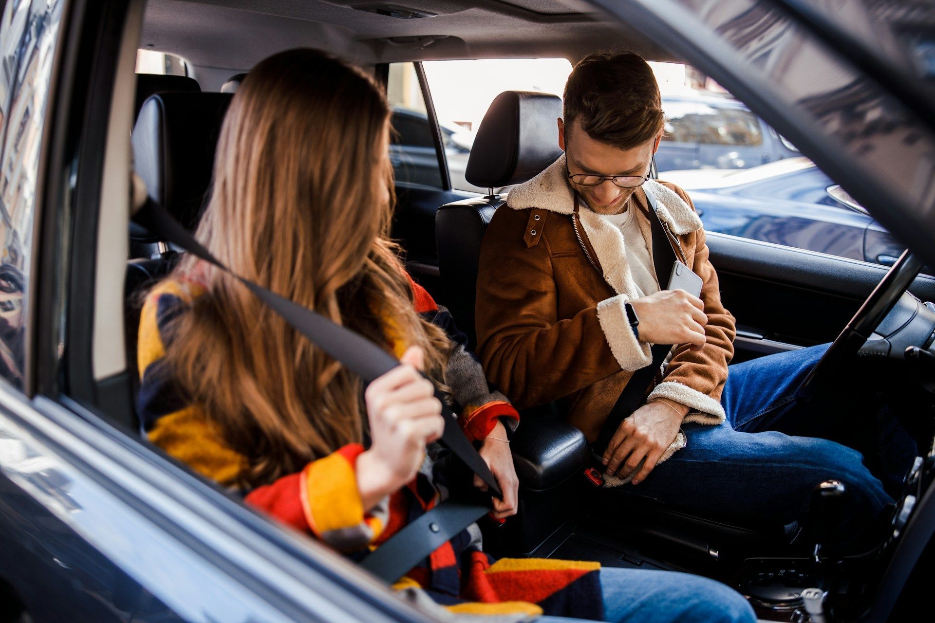 Woman and man inside a car fastening seatbelts. Man wears glasses and a brown jacket. Woman has long hair and a colorful jacket.