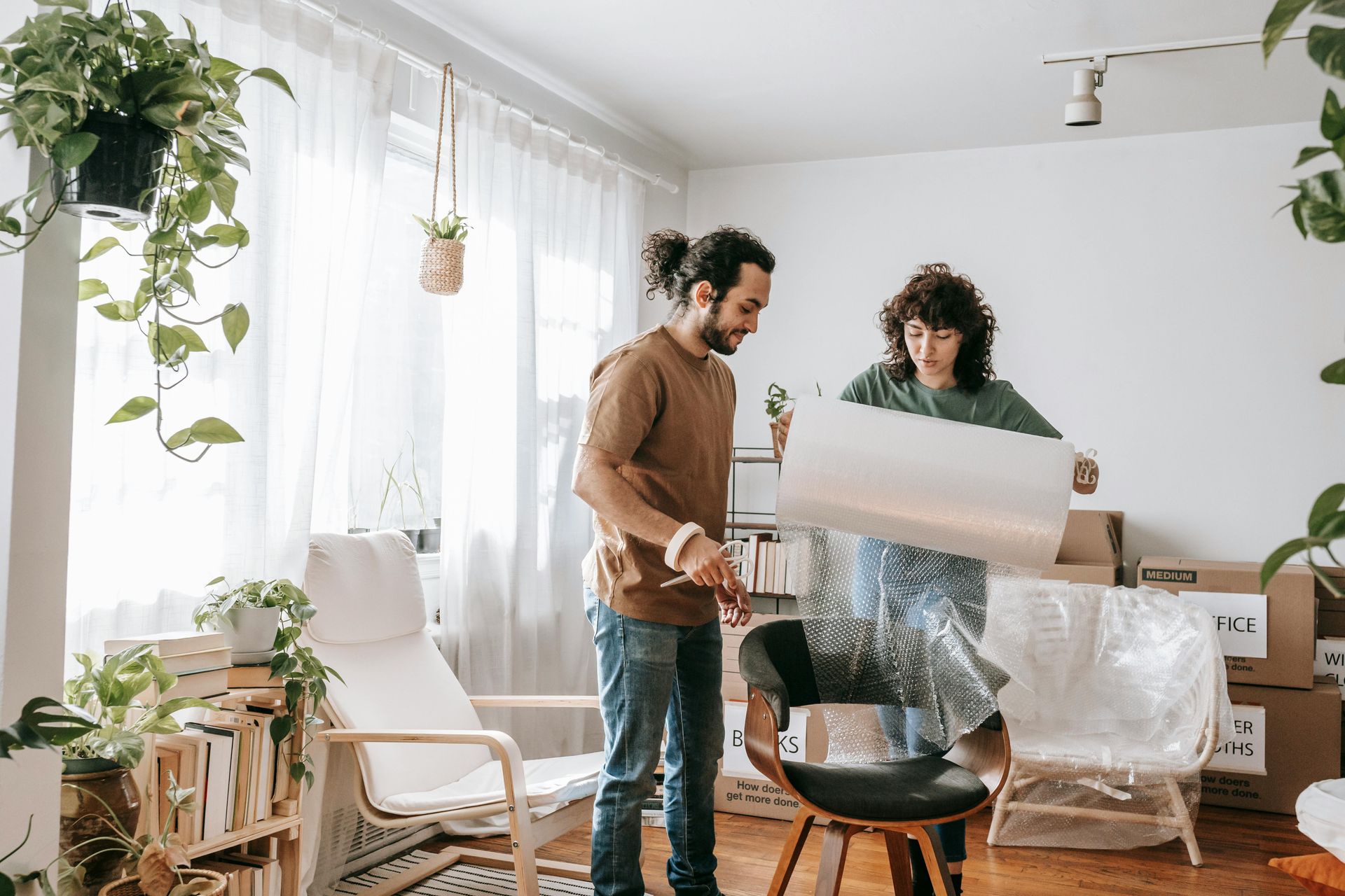 Couple wrapping chair in bubble wrap, preparing to move in a light-filled room with plants.