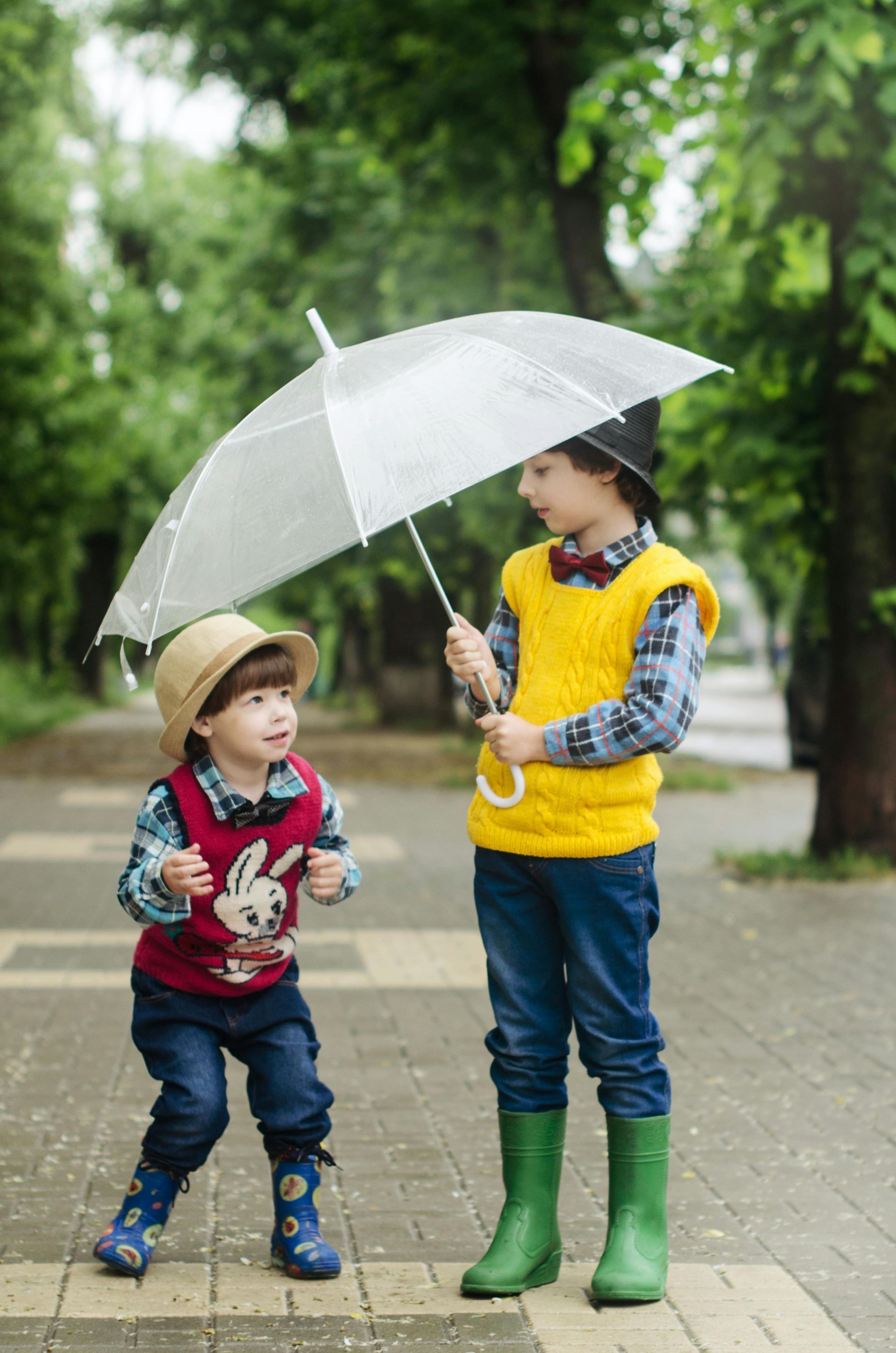 Two children in rain gear under an umbrella on a sidewalk. One child is holding the umbrella.