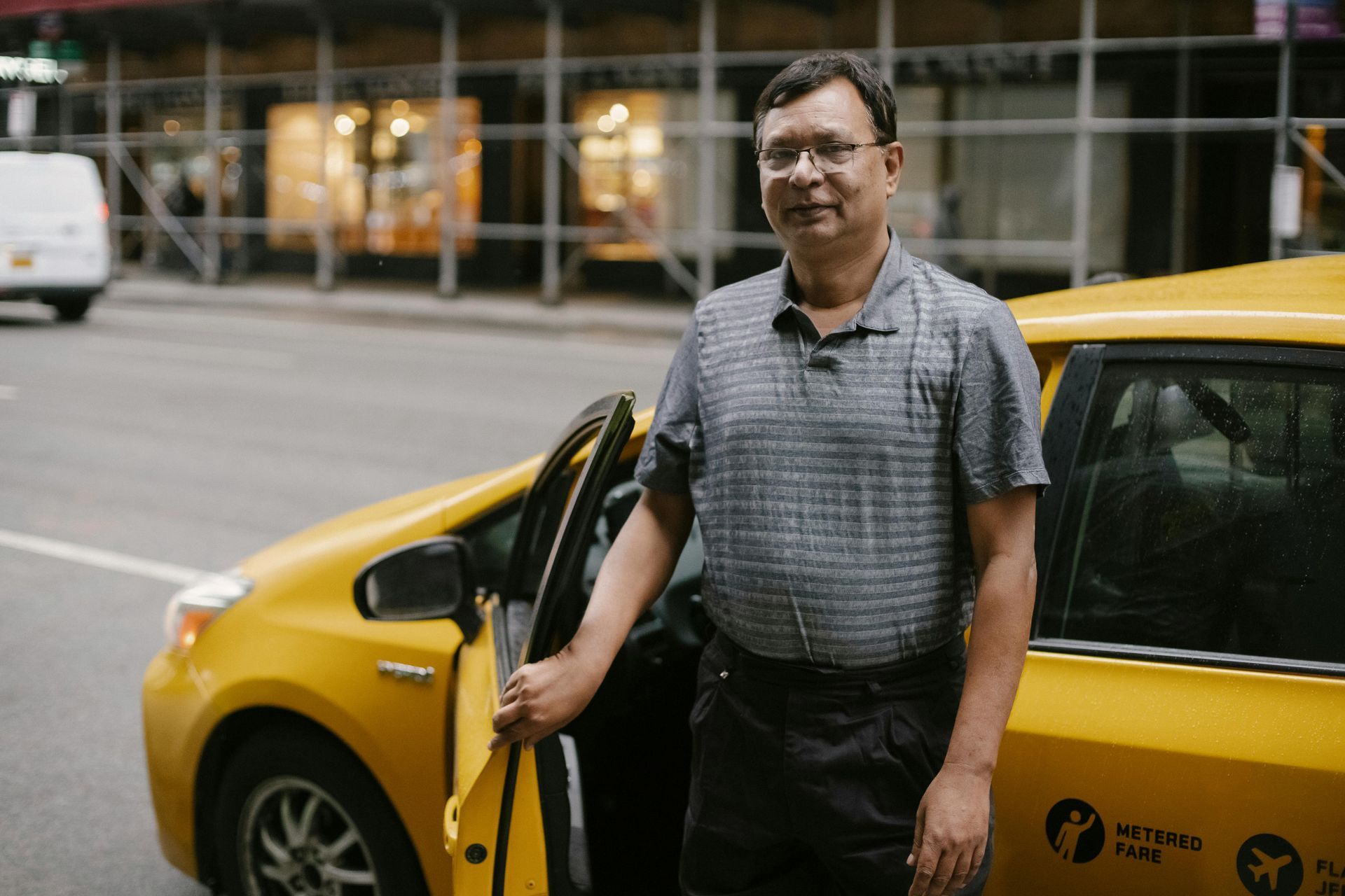 Man standing by a yellow taxi, holding the open door on a city street.
