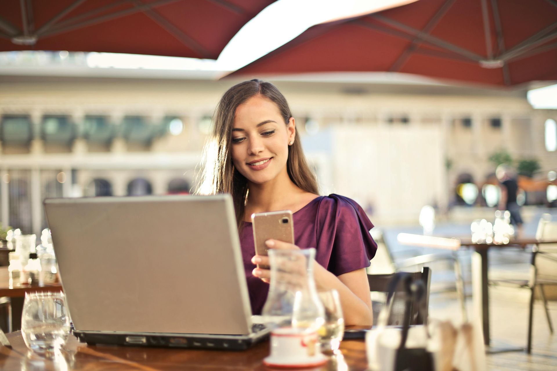 Woman sitting outdoors, using laptop and phone, smiling at phone screen.