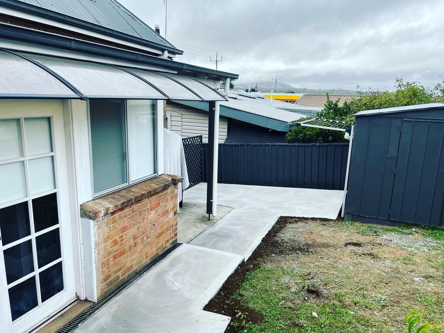 A gray concrete patio next to a brick wall with a shed and black fence under an overcast sky — LD Carpentry in Goulburn, NSW