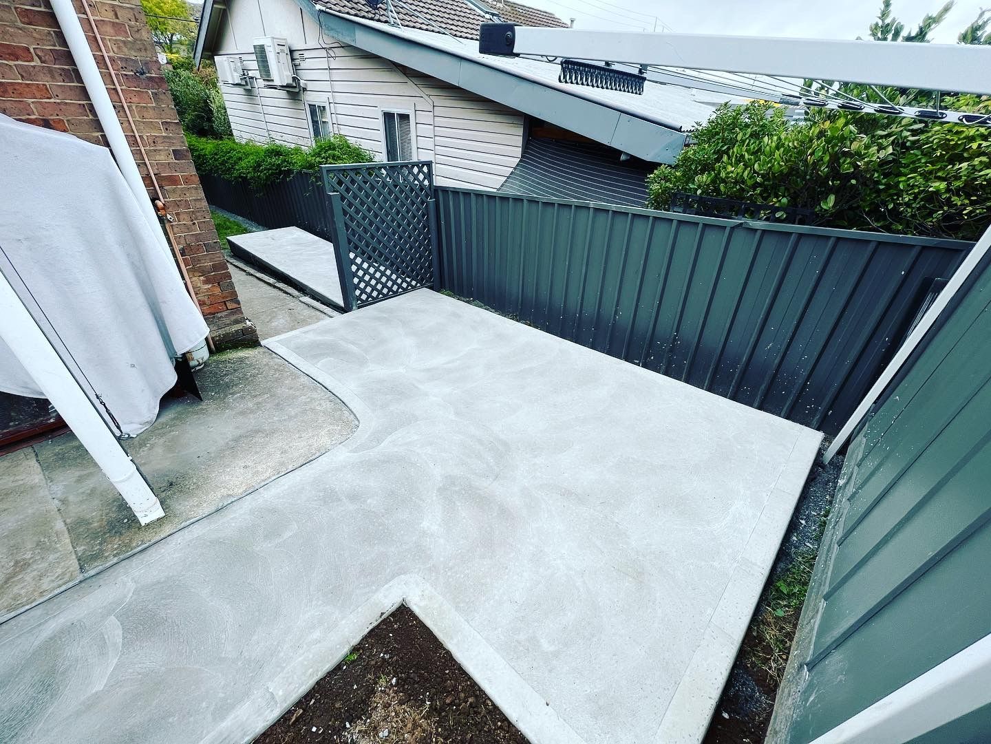 Concrete patio in backyard, bordered by gray fence and greenery, with a dark metal gate — LD Carpentry in Goulburn, NSW