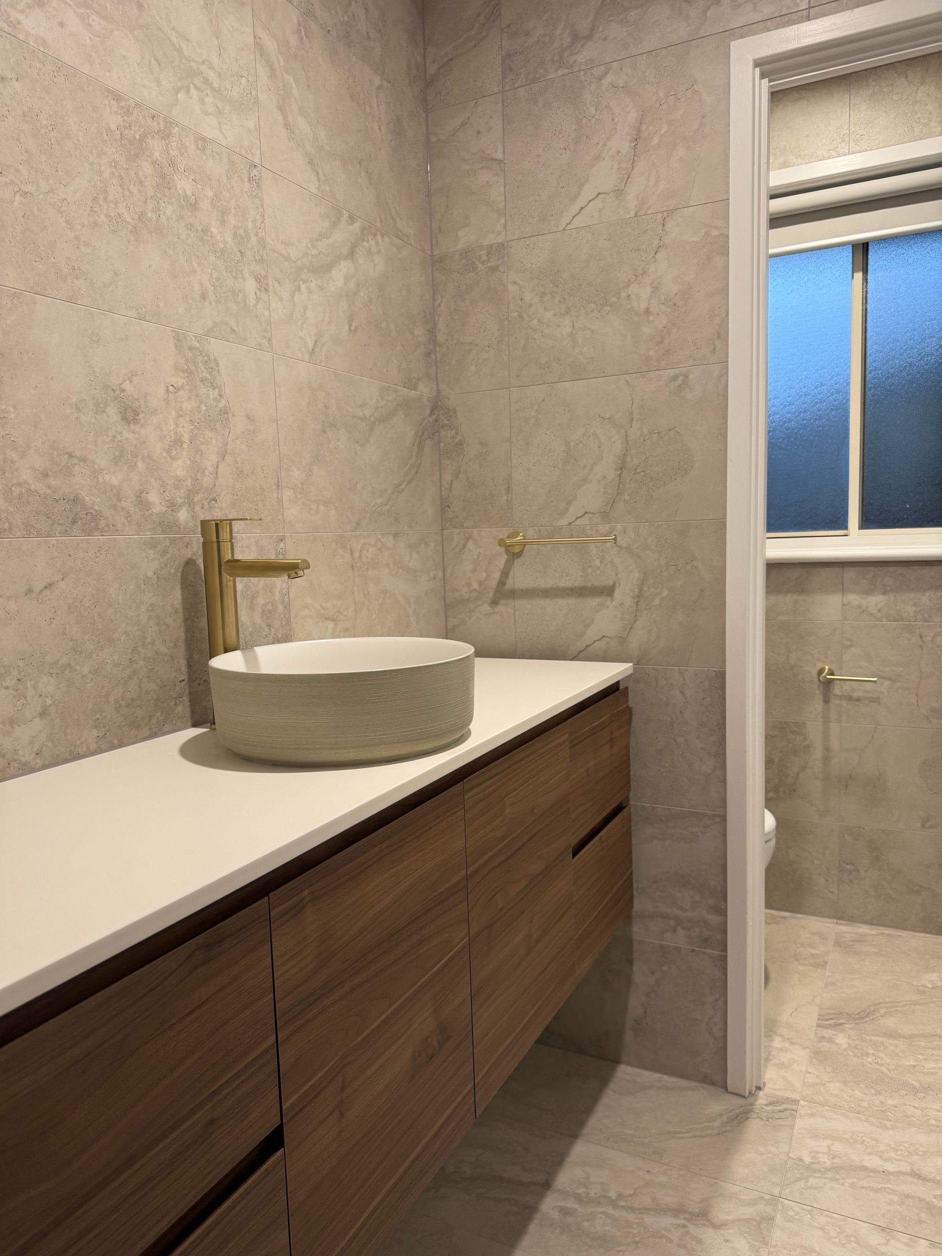 Bathroom with wood vanity, white countertop, gold faucet, and stone-like tile walls — LD Carpentry in Goulburn, NSW