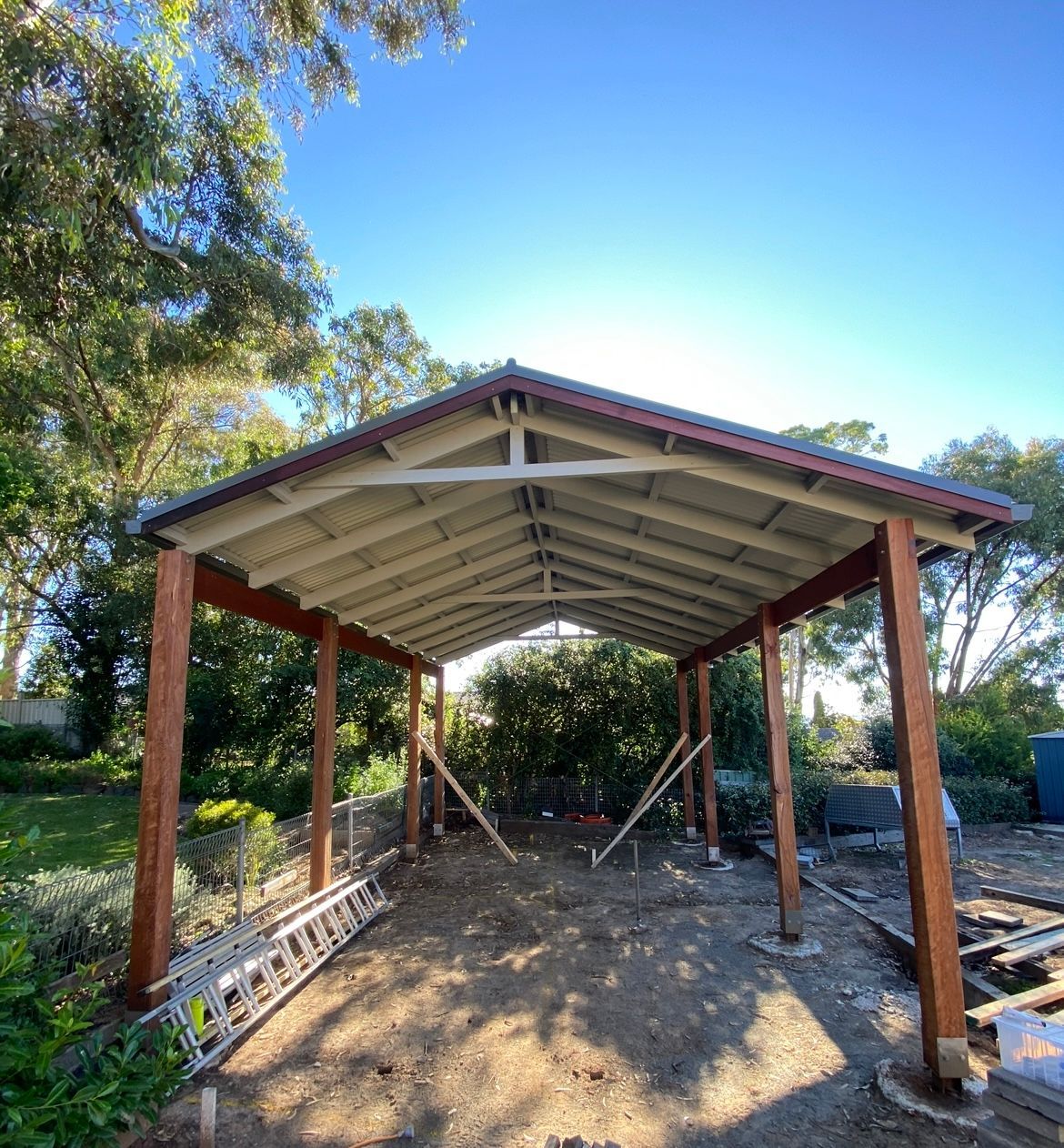 Carport under construction, brown wooden beams and supports, white roof, dirt ground, blue sky.