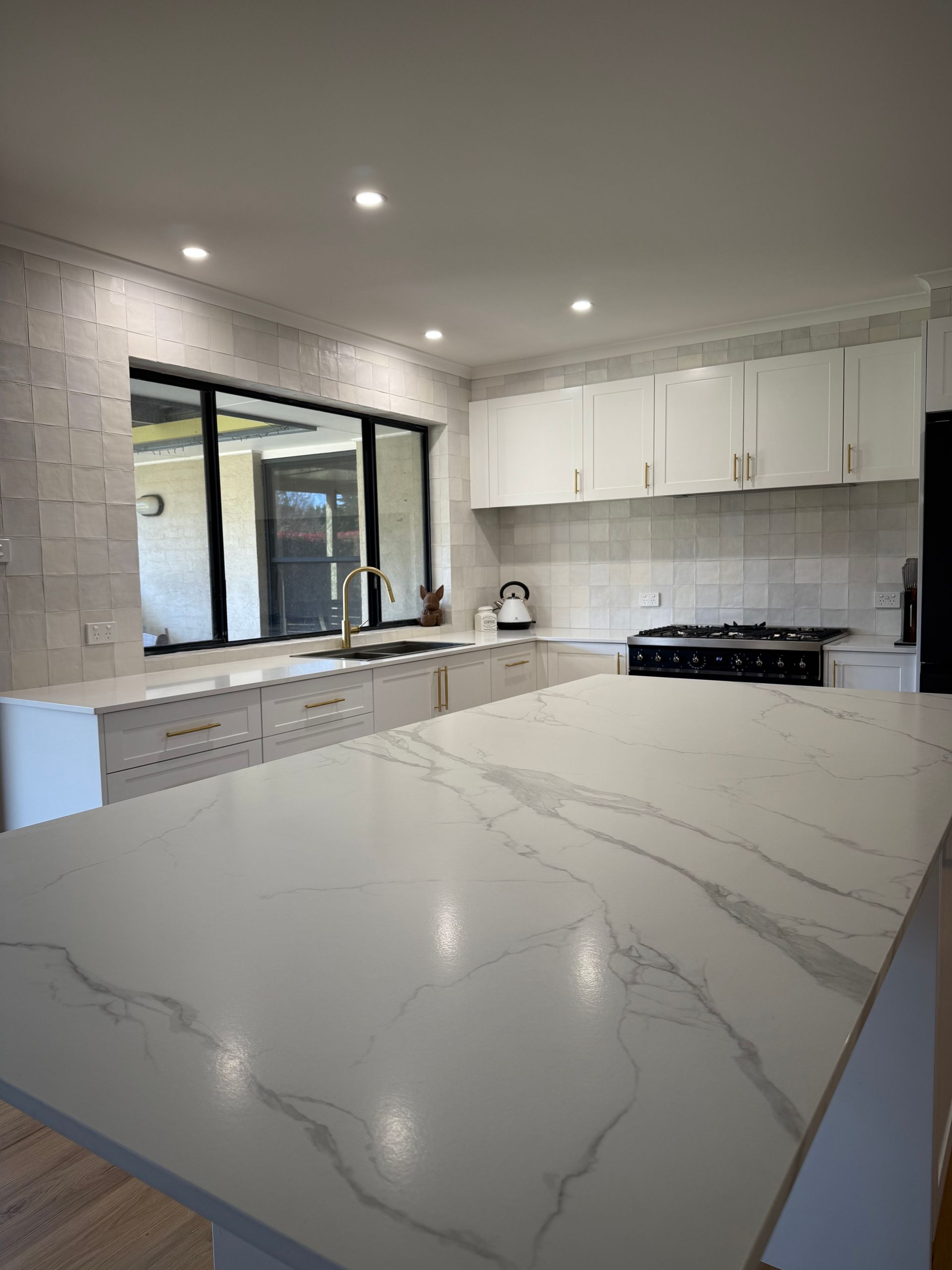 Modern kitchen with white cabinetry, countertops, and island. A large window and black stovetop are also visible.
