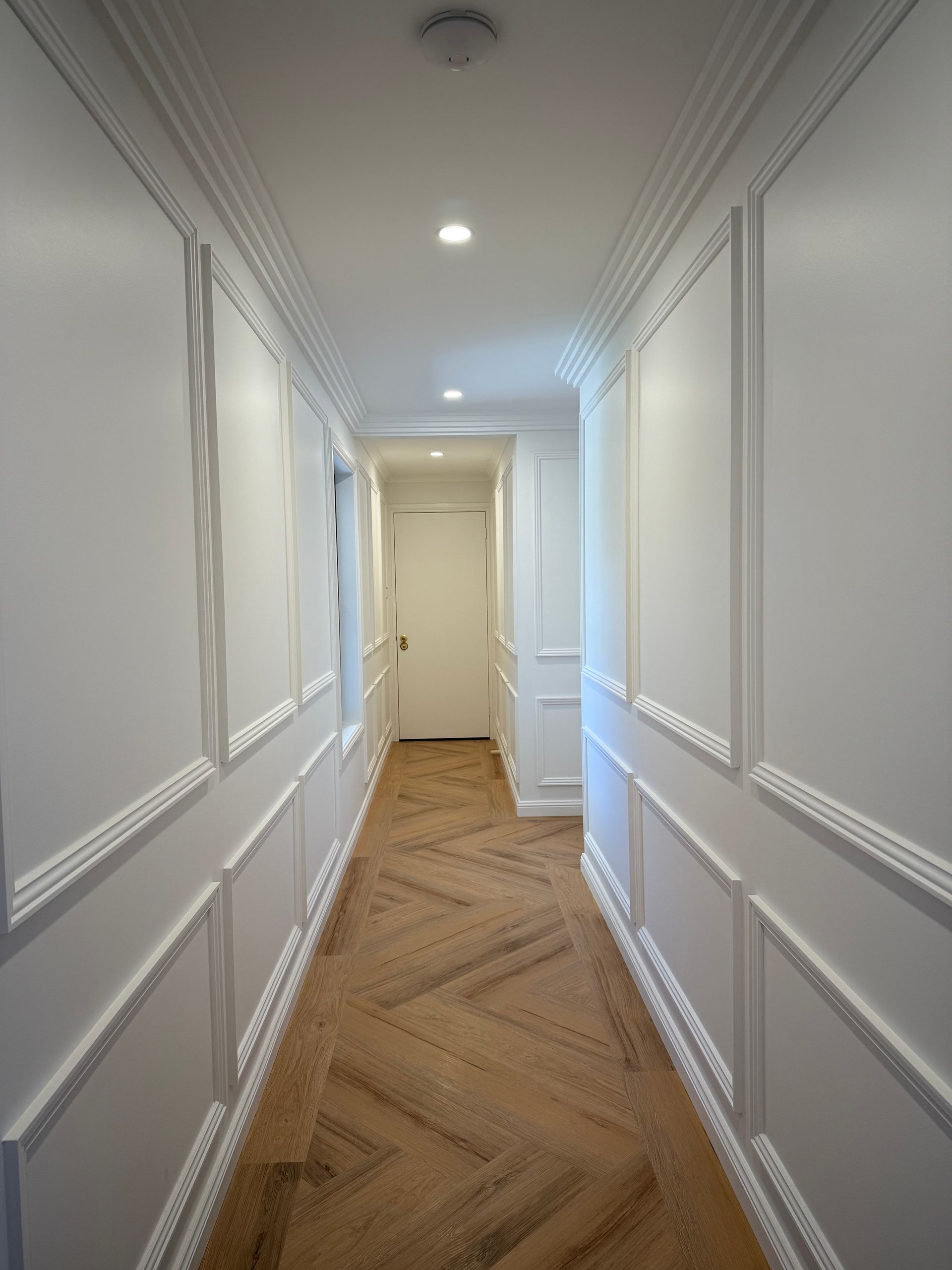 Narrow hallway with white panelled walls, ornate molding, wood floor, and a closed door at the end.
