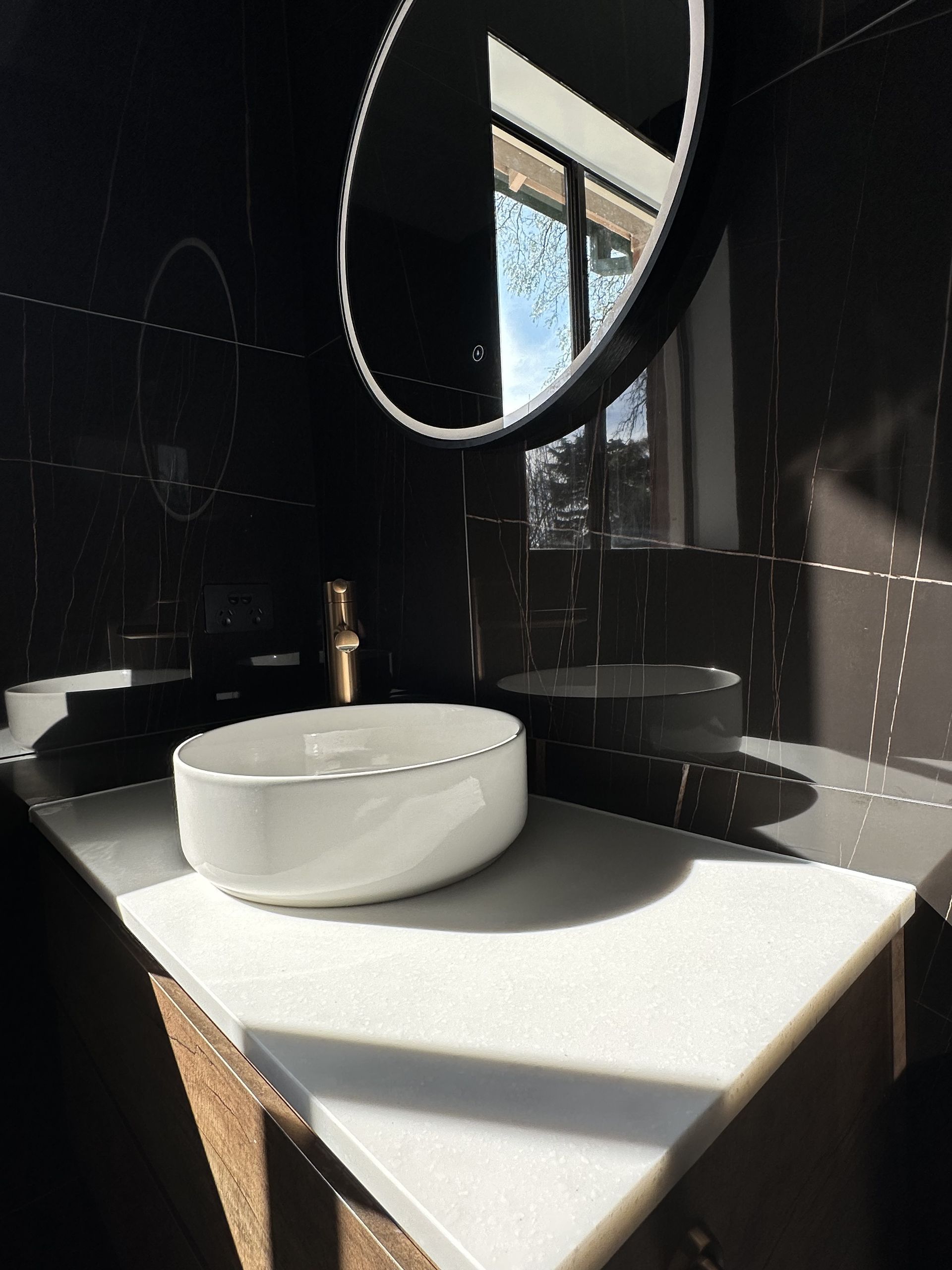 Bathroom with white countertop, black tile, round mirror, and white sink — LD Carpentry in Goulburn, NSW