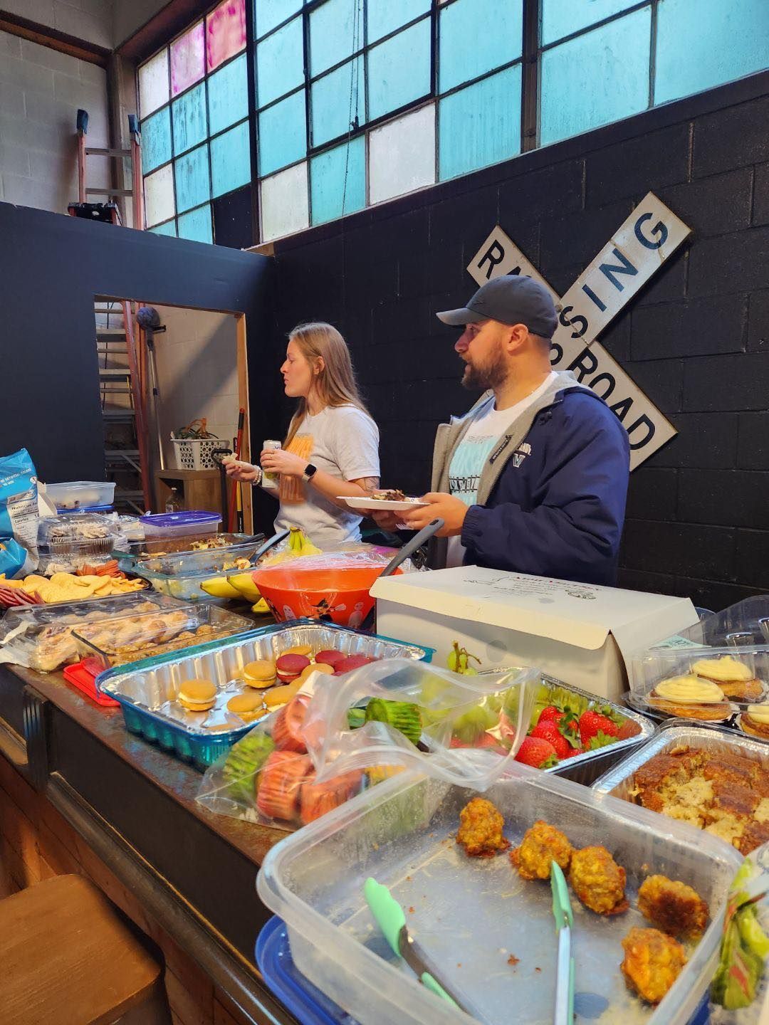 A man and a woman are serving food at a table with a railroad crossing sign in the background