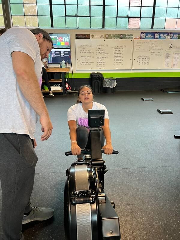 A man is standing next to a girl on a rowing machine in a gym.