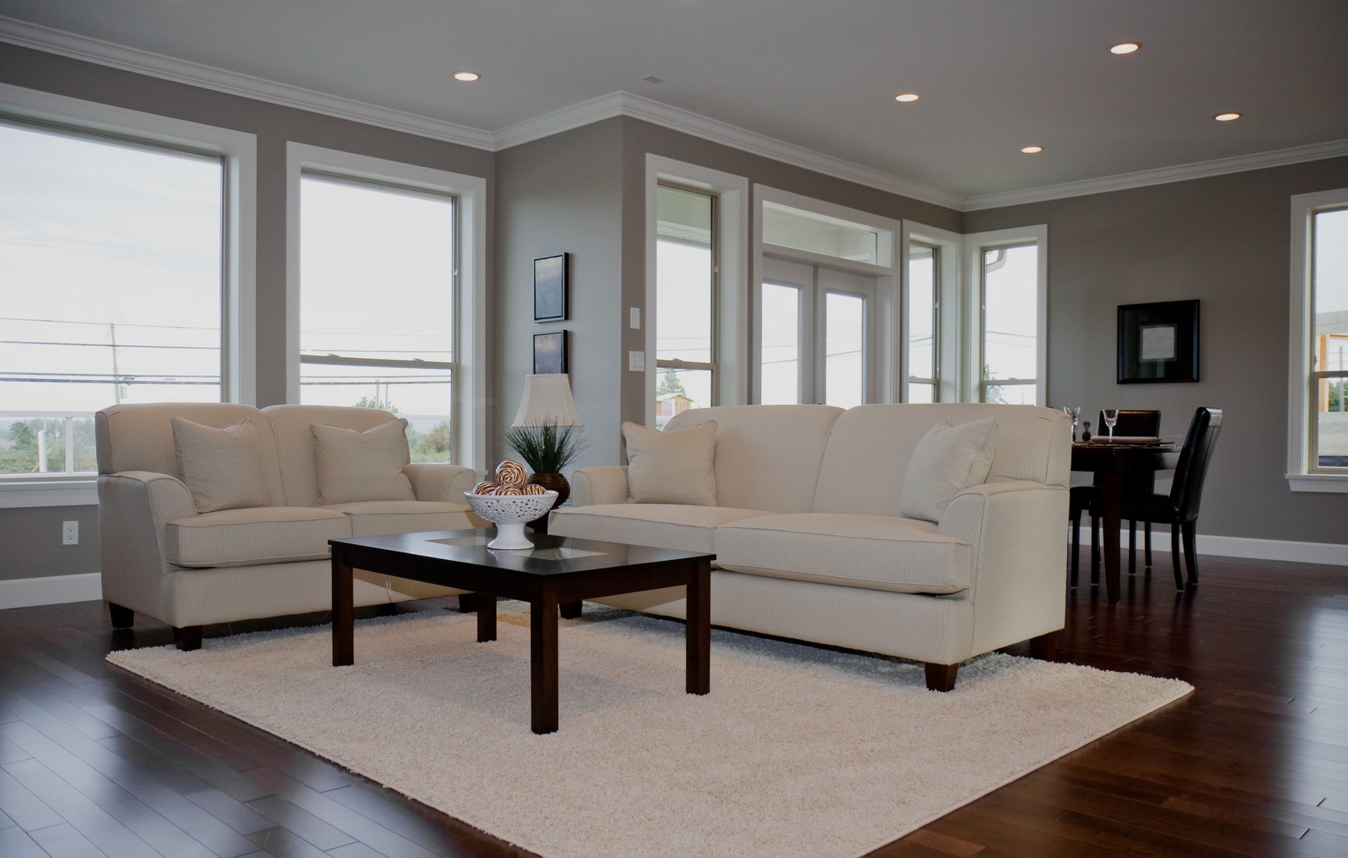 Living room with white couches, dark wood coffee table, and large windows; neutral walls and dark wood floors.