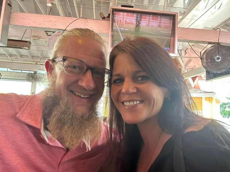 Man with a long beard and woman smile for a selfie. They are indoors with a bright background.