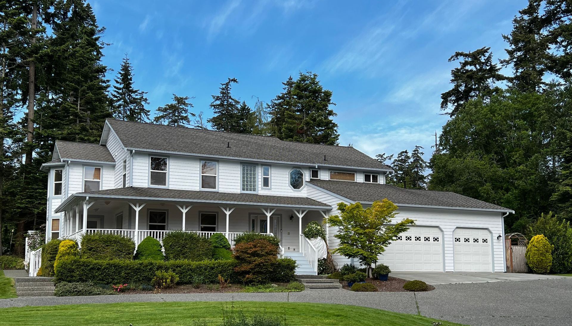 White two-story house with covered porch, attached garage, and blue sky background. Green trees and shrubs surround.