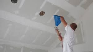 Man on ladder using drywall hopper to apply joint compound to ceiling.