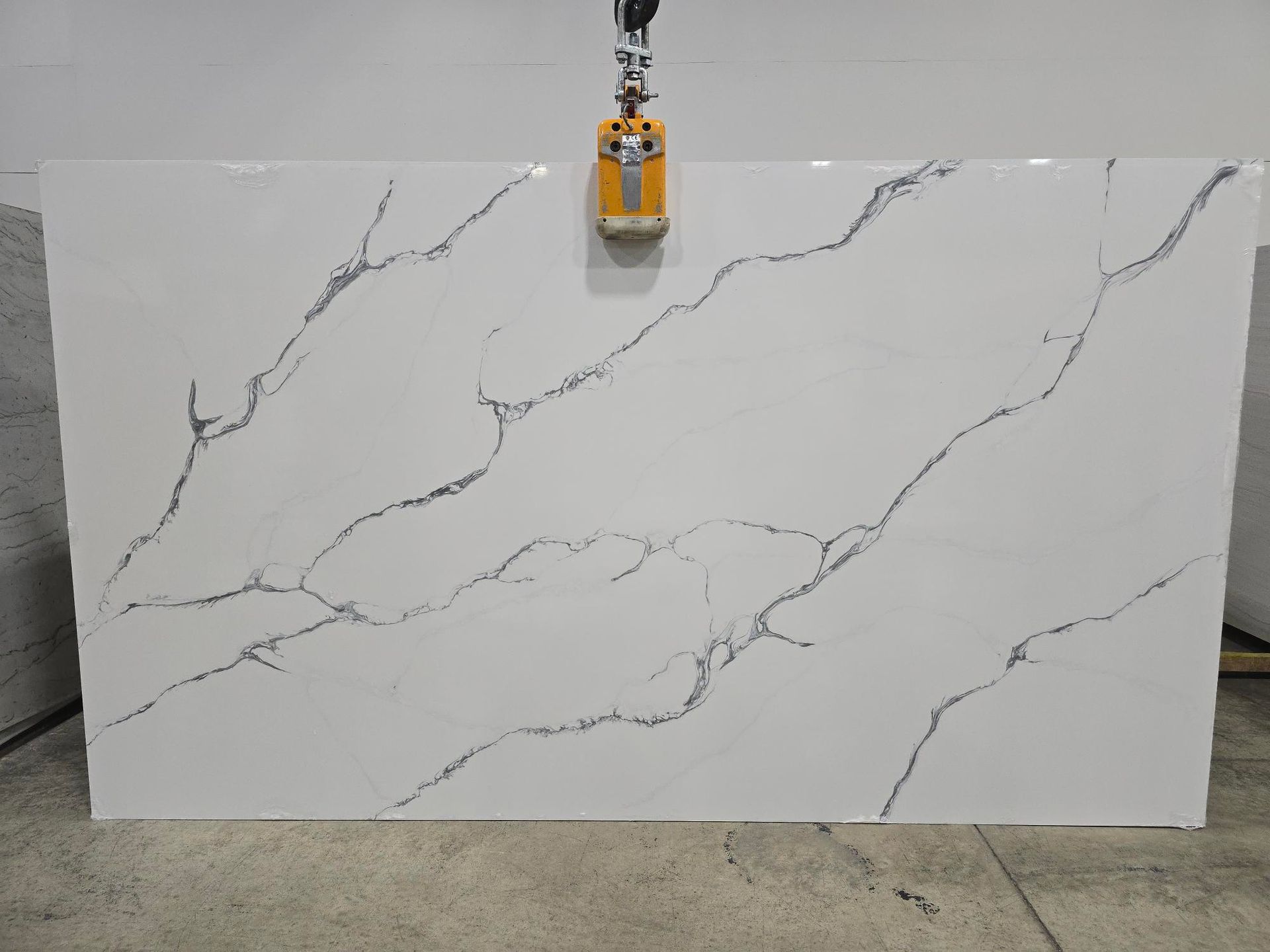 Large white marble-look slab with gray veining, hanging in a workshop by a clamp at the top center.