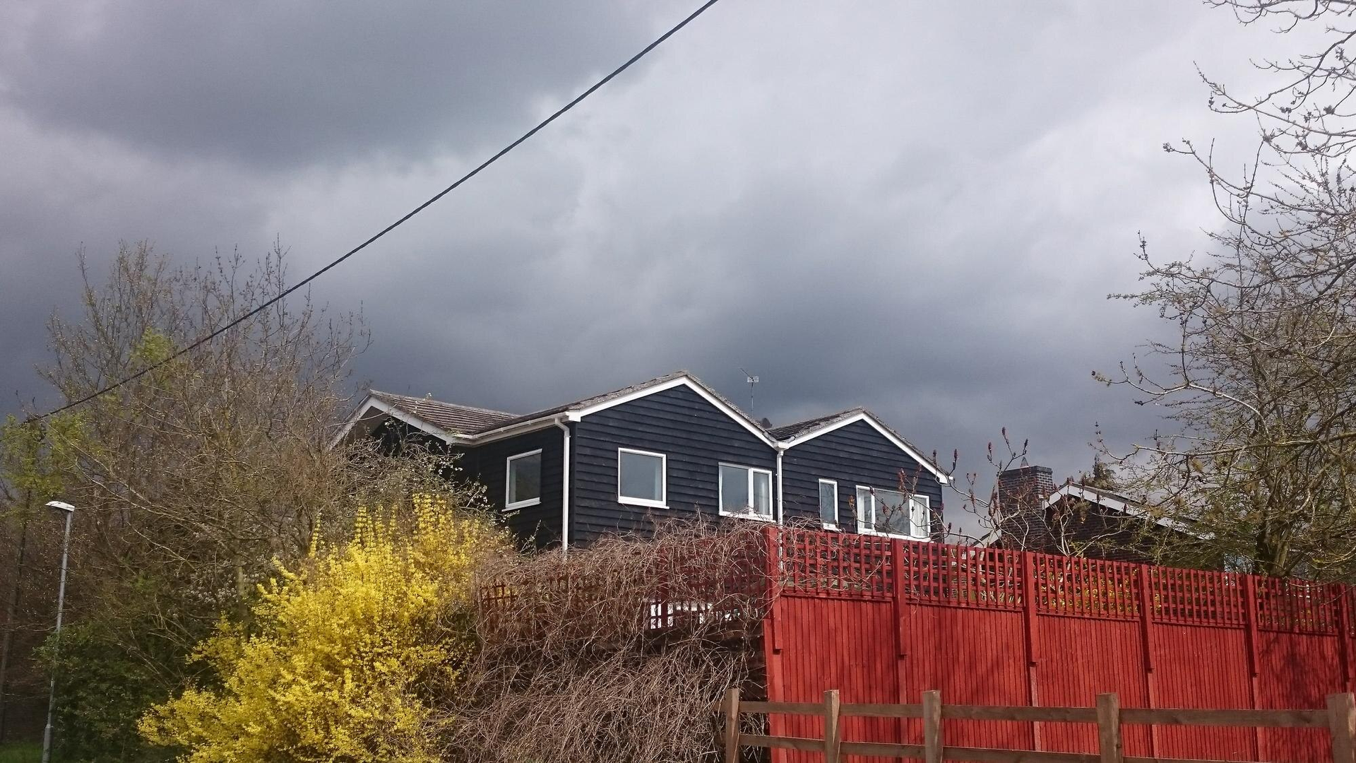 Dark house with white trim, red fence, and yellow and brown bushes under a cloudy sky.