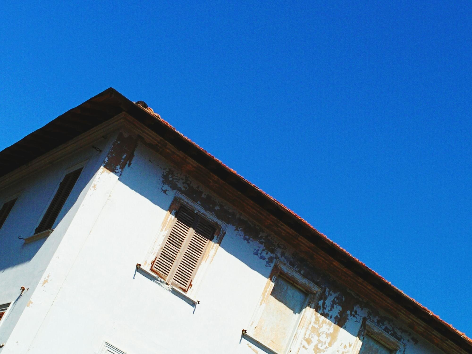 White building with peeling paint, brown roof against bright blue sky.