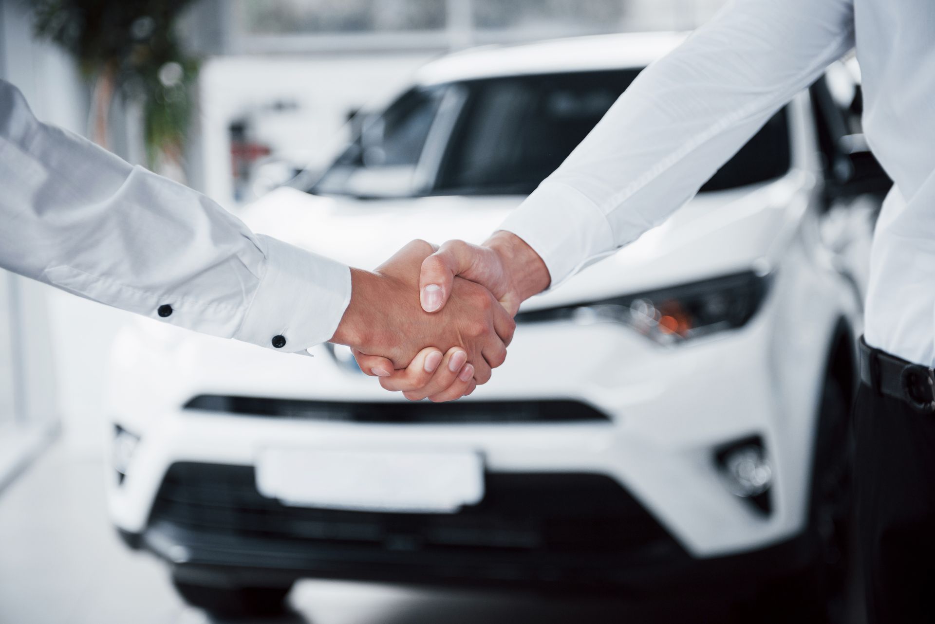 Two men are shaking hands in front of a white car.