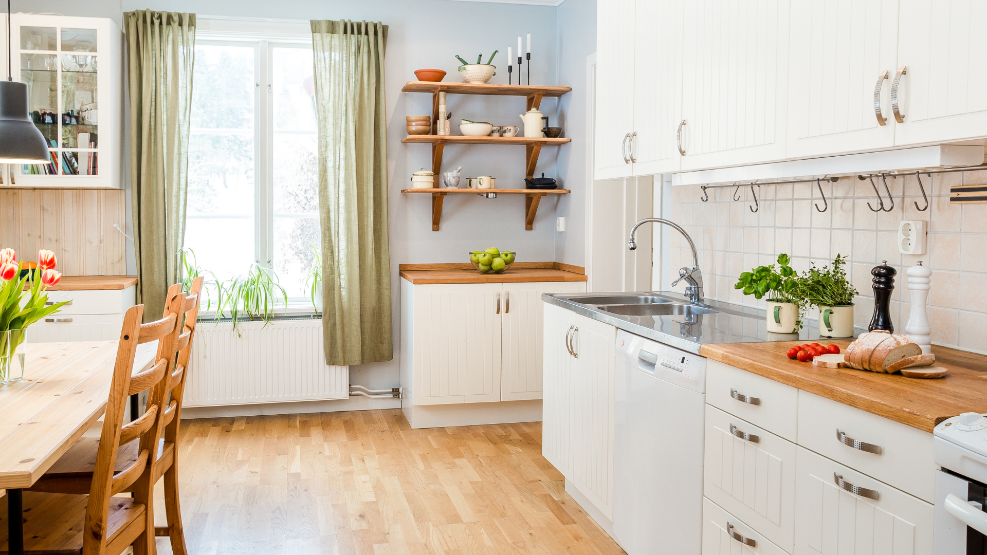 A bright, airy kitchen with white cabinetry, light wood countertops, open shelving, and a dining area with green curtains.