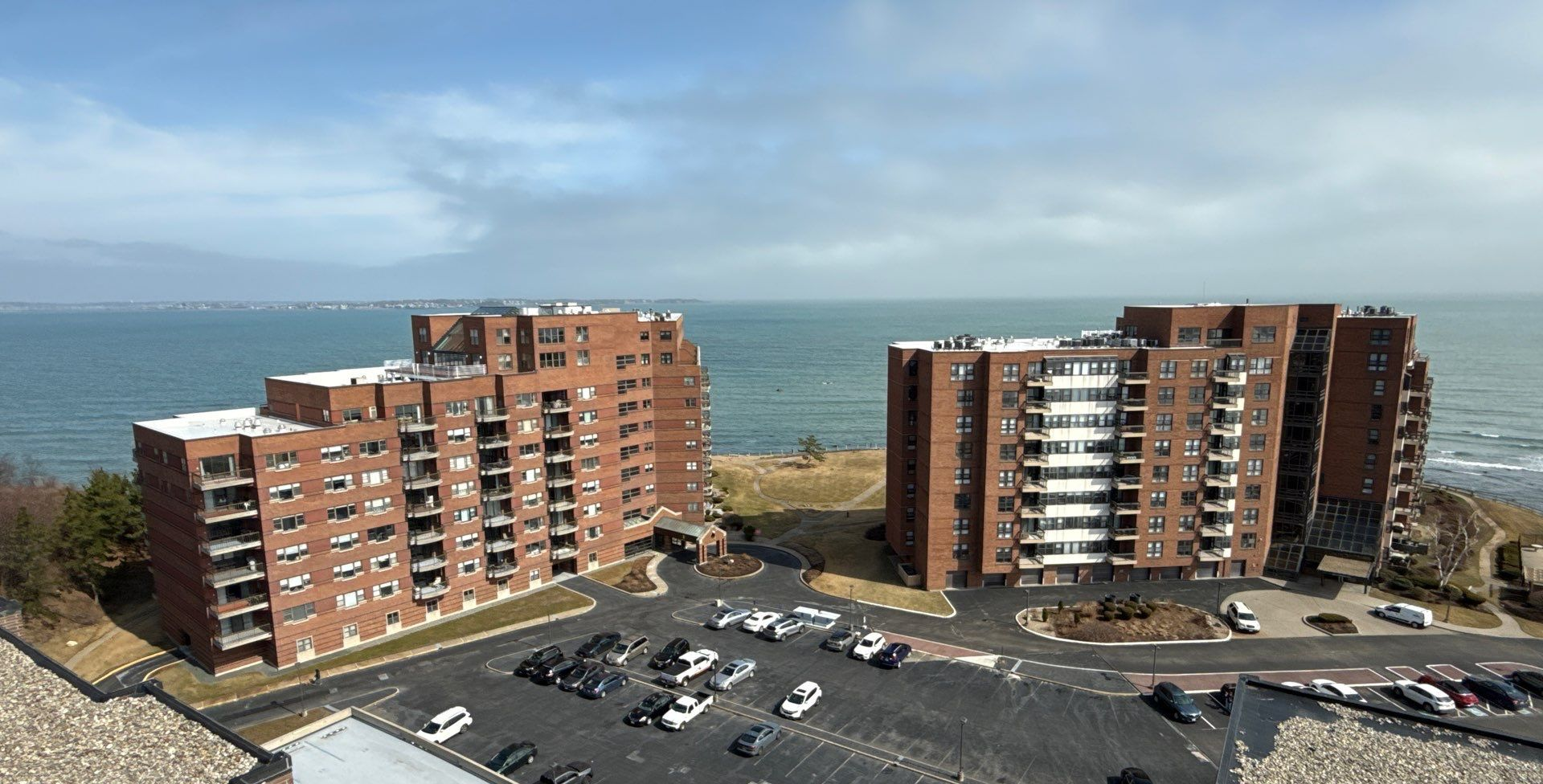 An aerial view of a multi-story apartment building with a dark shingled roof, surrounded by parking and lawns.