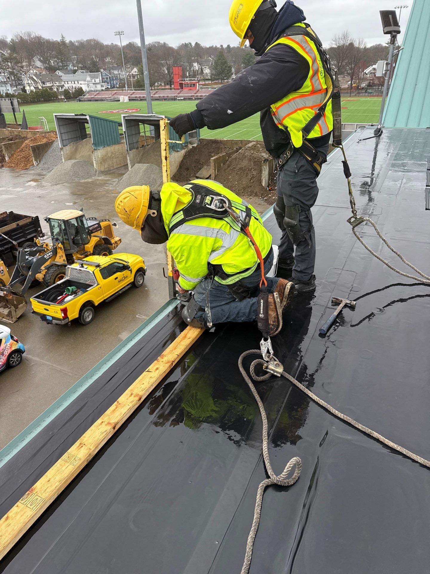 Two construction workers in high-visibility safety gear work on a roof edge, measuring a wooden board near a yellow vehicle.