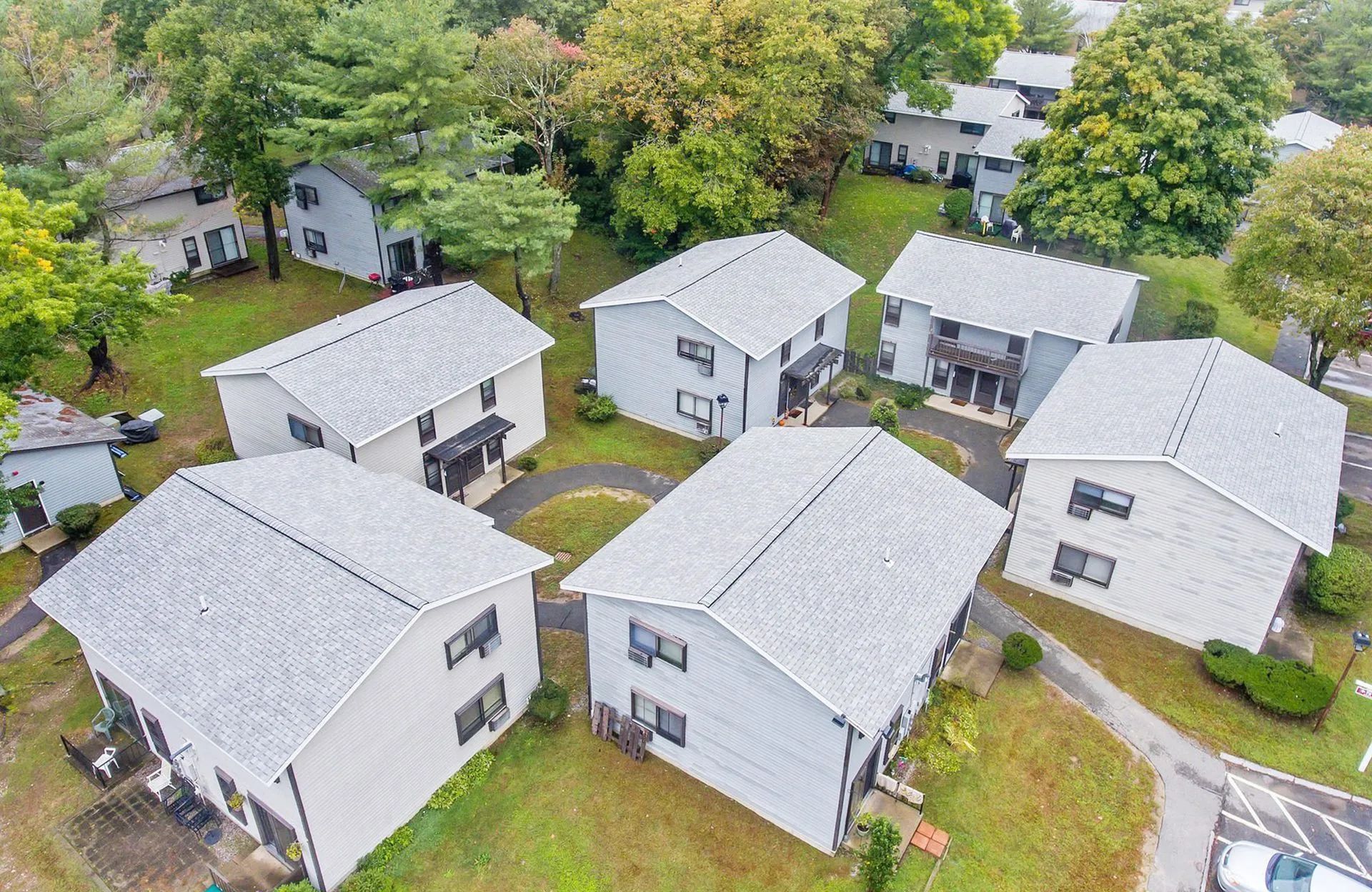 Aerial view of several gray houses with light-colored roofs, surrounded by green grass and trees.