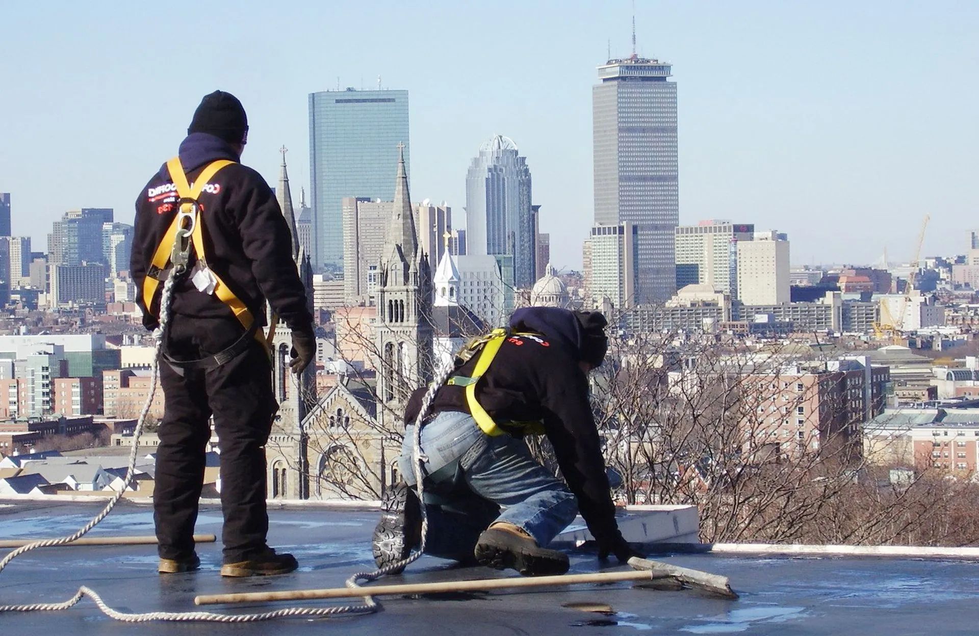 Two roofers wearing harnesses on a rooftop in a city, with buildings and a bright sky visible.