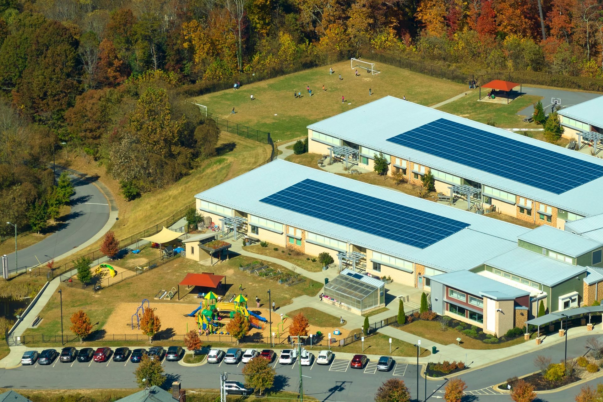 Aerial view of a school building with solar panels on the roof, surrounded by a playground, parking lot, and trees.