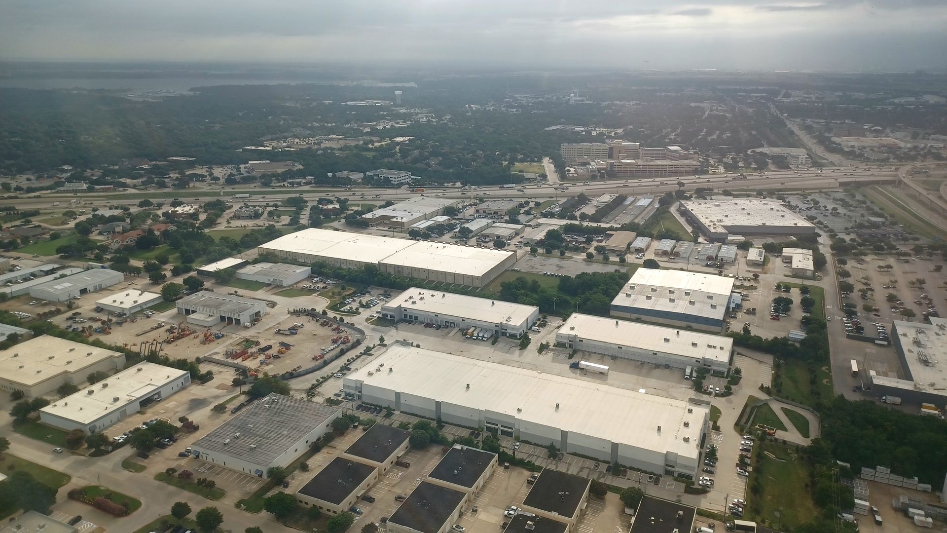 Aerial view of several flat-roofed warehouse and industrial buildings surrounded by parking lots and a nearby city.