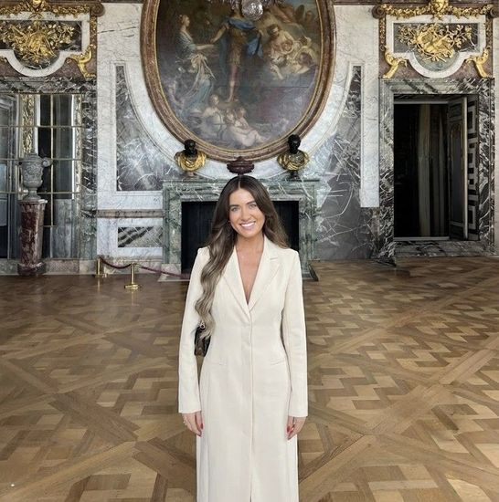 Woman in a white dress stands in a grand, ornate room with a chandelier and large painting.