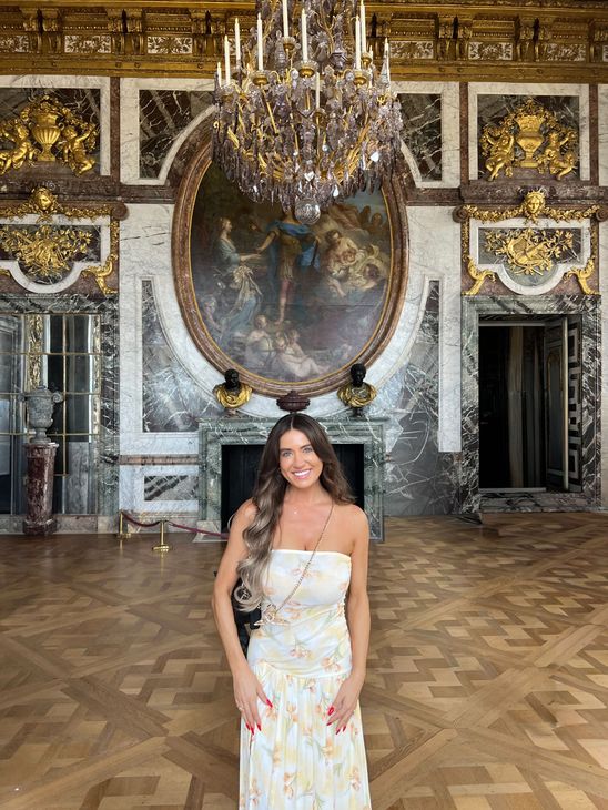Woman in a white dress stands in a grand, ornate room with a chandelier and large painting.