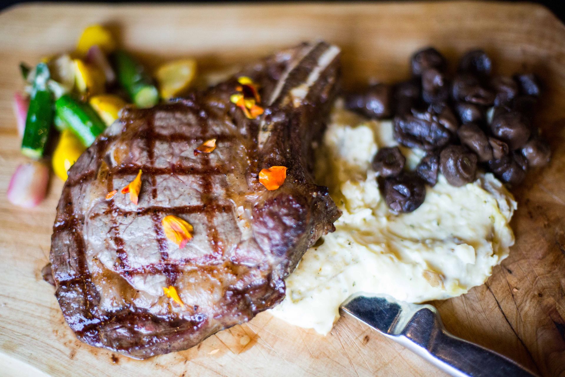A steak with mashed potatoes and vegetables on a wooden cutting board.