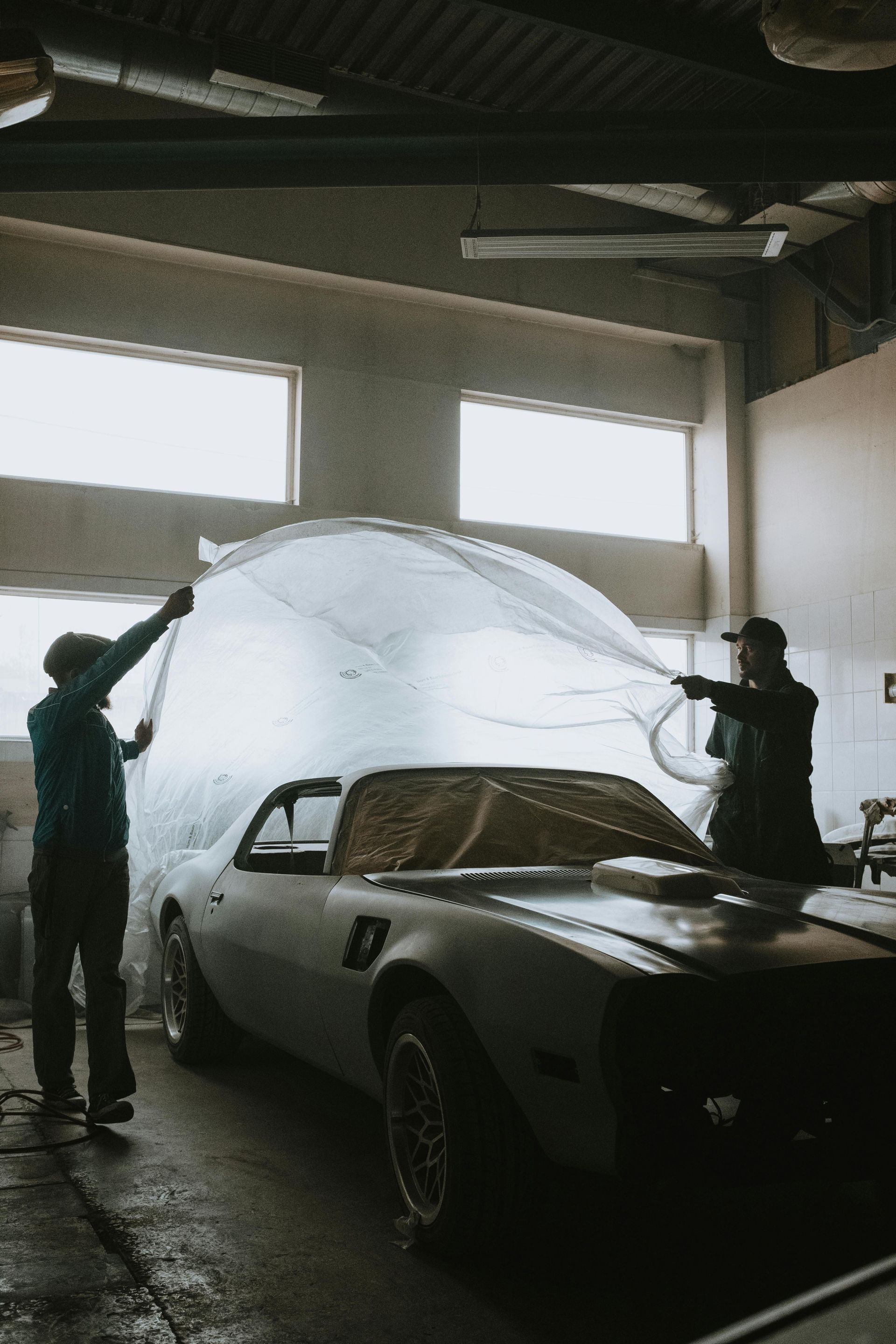 Two people covering a silver classic car with a large plastic sheet in a garage.