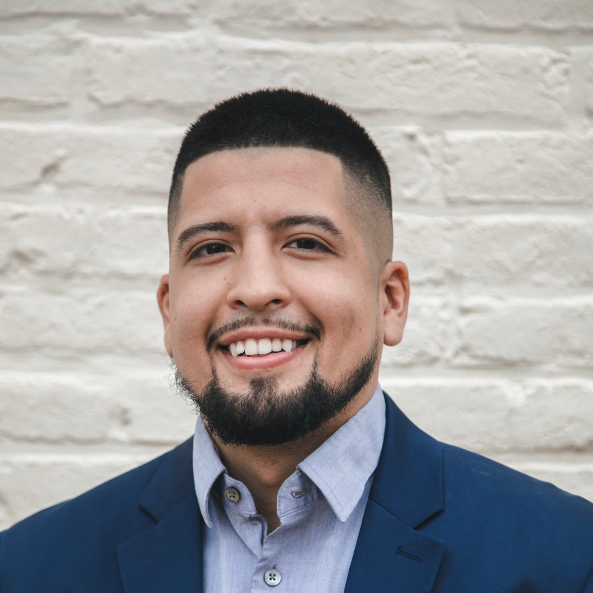 A man with a beard and a blue suit is smiling in front of a white brick wall.