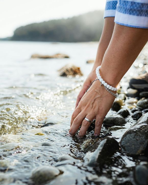 Eine Frau berührt am Strand Steine im Wasser und trägt ein Steinarmband und Silberschmuck.