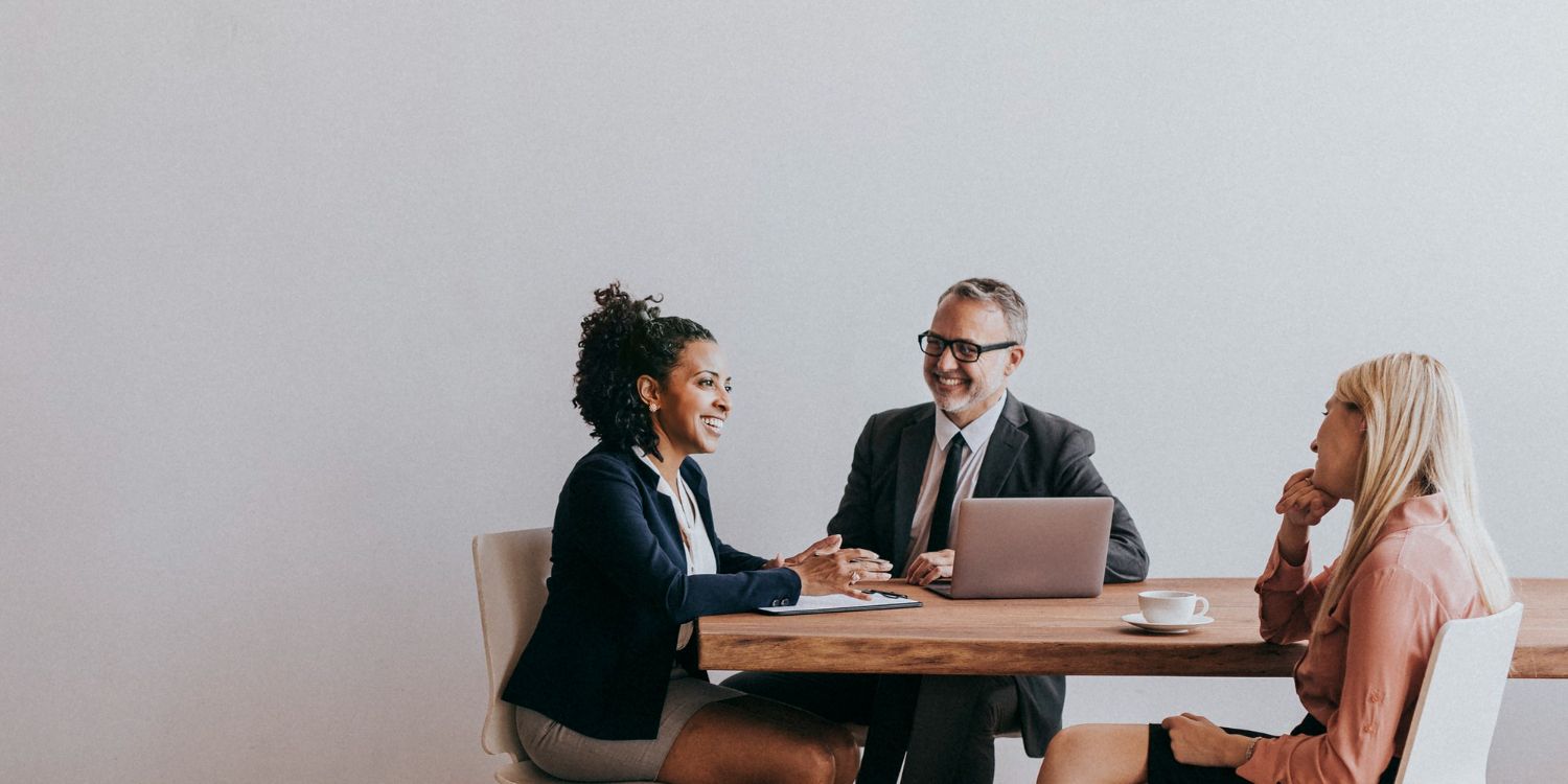 Group of people in a bright office, discussing papers. A woman holds documents, others listen.