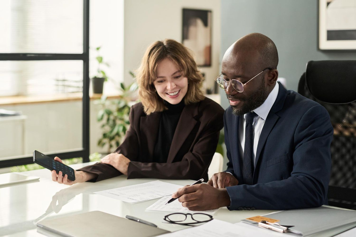 Group of people in a bright office, discussing papers. A woman holds documents, others listen.