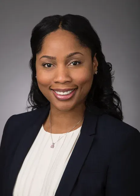 Woman in a navy blazer and white top smiles, looking at the camera against a gray backdrop.