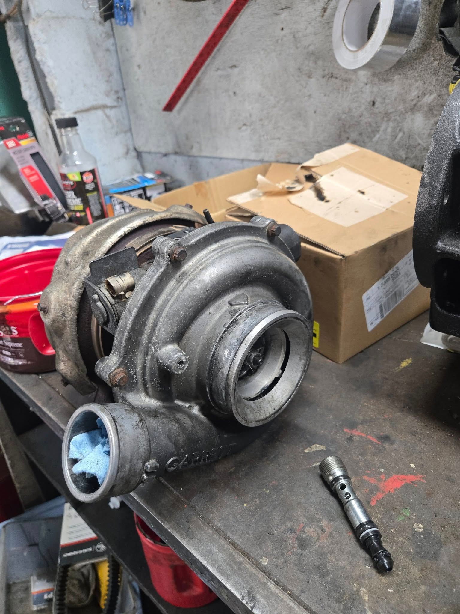 A disassembled turbocharger on a workbench in a cluttered garage, with a box and tools in the background.