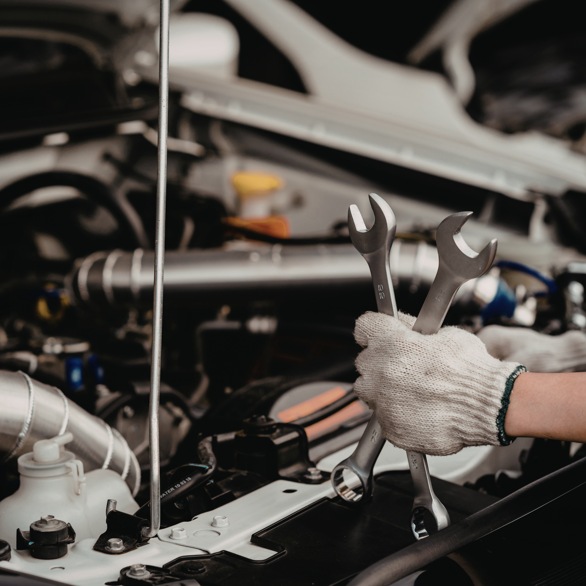 Gloved hand holding wrenches over an open car hood, with engine components visible.