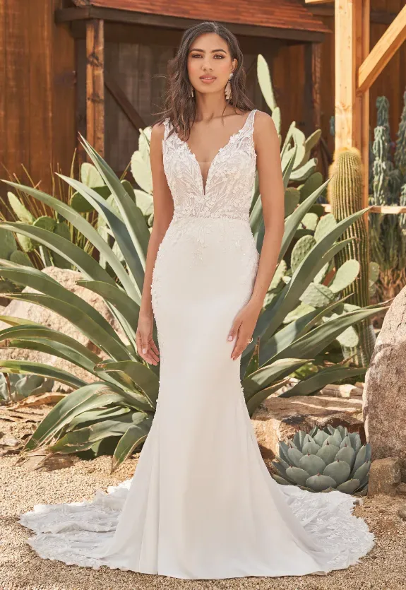 Woman in white wedding dress, posing outdoors with plants, building in background.