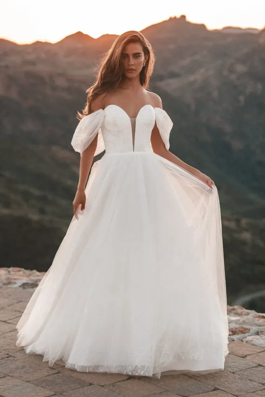 Woman in a white off-shoulder wedding dress poses on a hilltop at sunset.