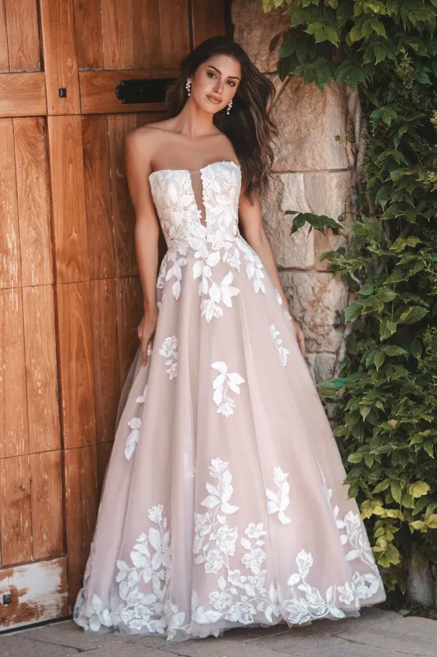 Woman in a strapless white and blush floral wedding gown leans against a wooden door.