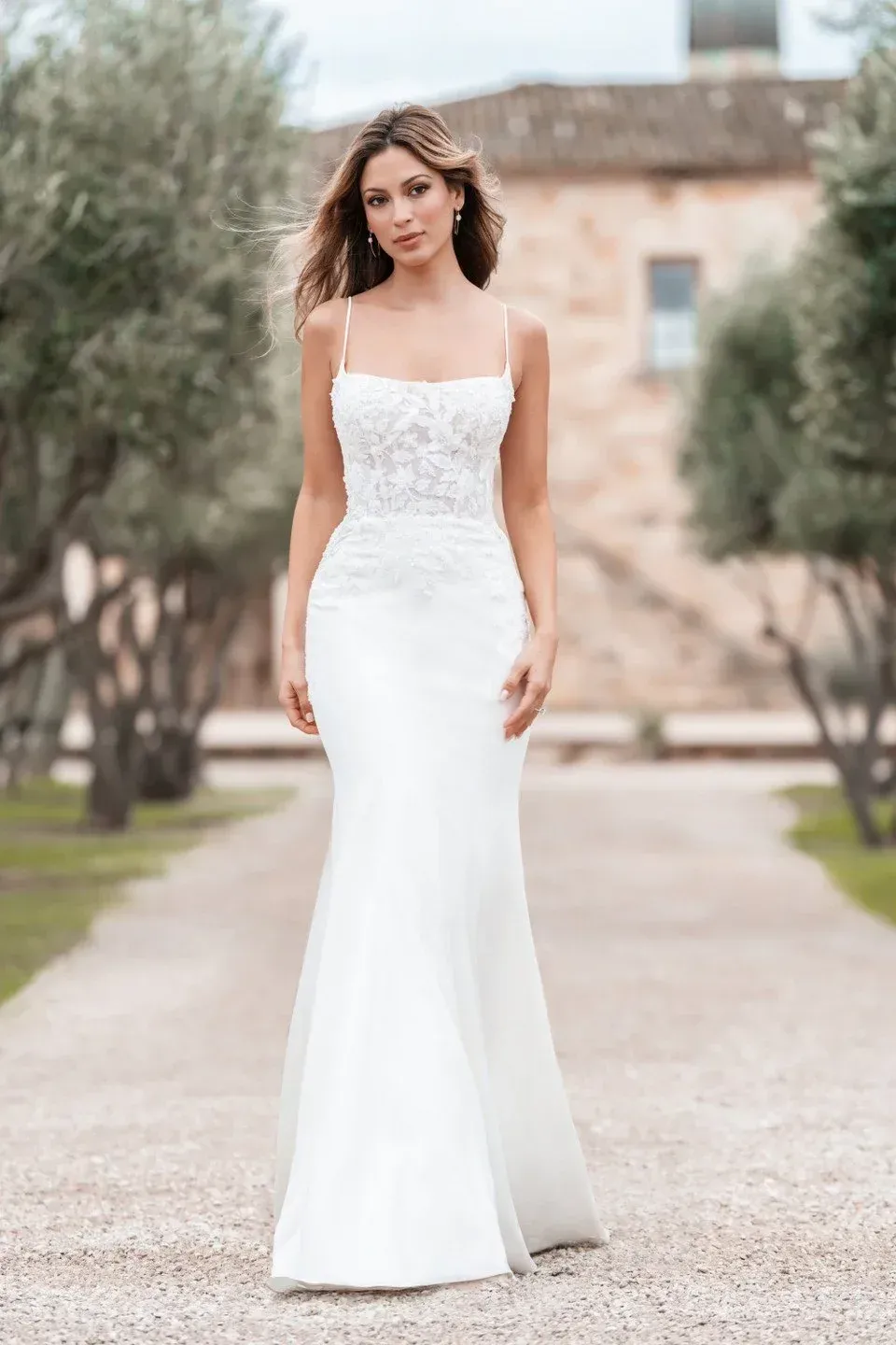 Woman in white wedding dress, spaghetti straps, lace bodice, posed outdoors on a pathway.