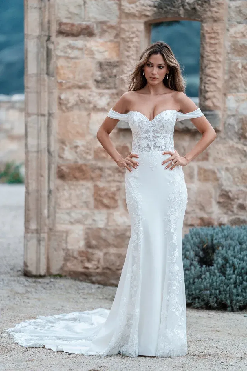 Woman in white off-the-shoulder wedding dress stands with hands on hips in front of a stone wall.